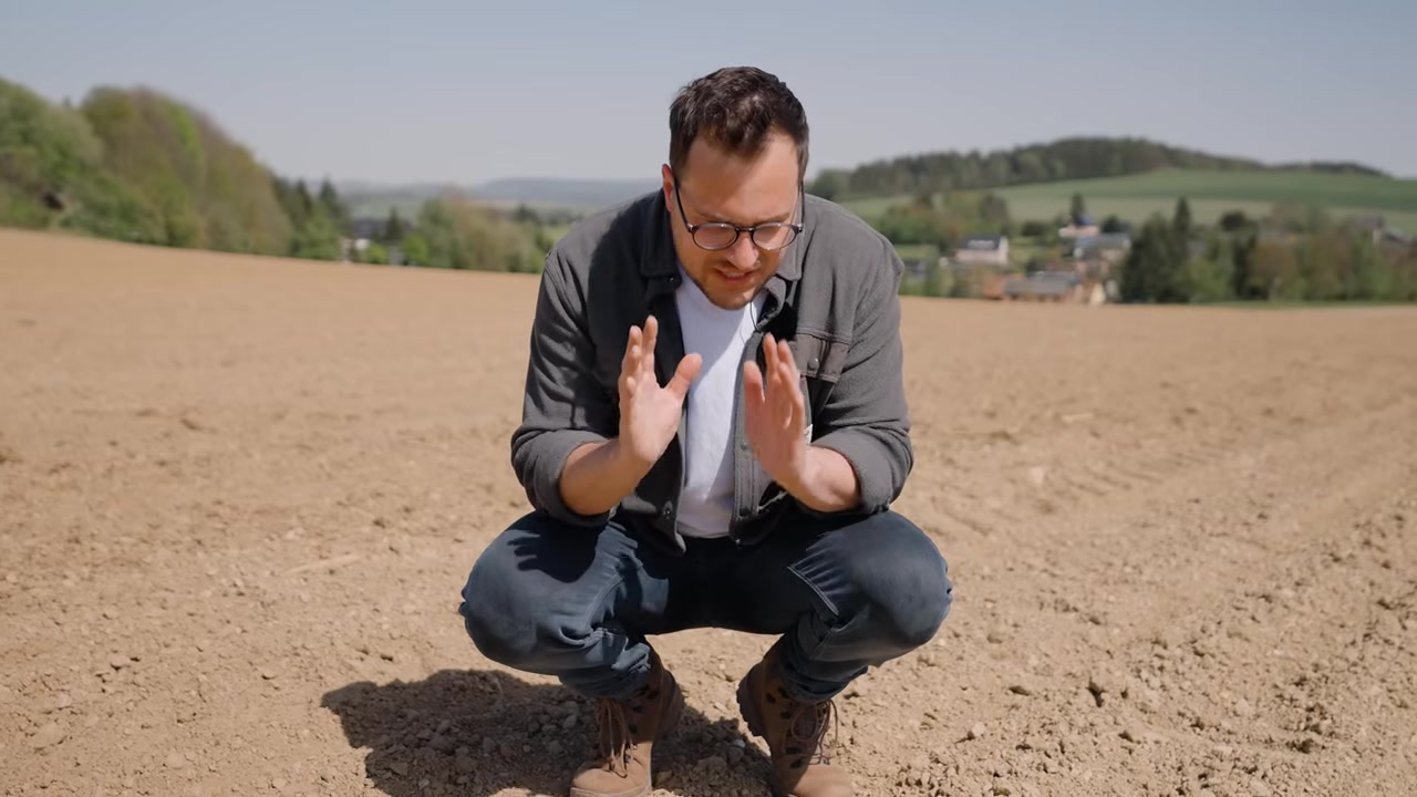 Moderator kniet auf dem frisch bestellten Feld und erklärt die Saatgut-Ablage